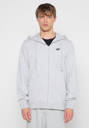 Młody mężczyzna ubrany w jasnoszarą bluzę z kapturem na zamek z sznurkami i czarne logo na piersi, stojący na jednolitym białym tle.