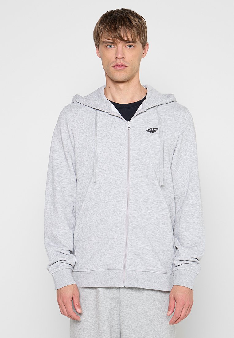 Jeune homme portant un sweat à capuche gris clair zippé avec cordons et logo noir sur la poitrine, debout devant un fond blanc uni.