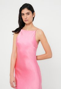 Ροζ σατέν φόρεμα με κομψό, εφαρμοστό σχέδιο και λεπτές τιράντες. Λείος υφασμάτινος με διακριτική λάμψη, ελάχιστες λεπτομέρειες και τονισμένες ραφές στα πλευρά.