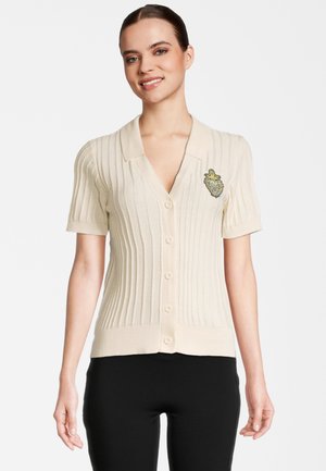 Kvinde iført en cremefarvet ribbet kortærmet cardigan med knapper og et dekorativt emblem på venstre bryst, parret med sorte bukser.