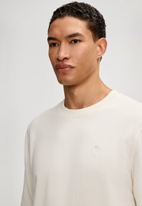 Jasna kremowa bluza z okrągłym dekoltem, wykonana z miękkiego materiału, z ribbowanym wykończeniem rękawów oraz małym haftowanym logo na piersi. Gładka tekstura.