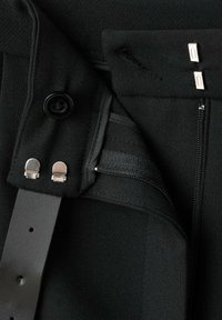 Zwarte broek heeft een ritszak, knoopsluiting en metalen accenten. De stof lijkt glad met een lichte textuur.