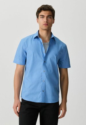 Young man wearing a short-sleeve light blue checkered button-up shirt over a gray t-shirt with black pants, standing against a plain background.