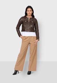 Giacca cropped in pelle marrone con due tasche frontali, indossata sopra una maglietta bianca e pantaloni a gamba larga beige, abbinata a stivali neri con tacco.