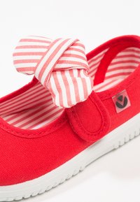 Zapato infantil de lona roja con suela blanca, con un lazo de tela a rayas rojas y blancas y forro interior a juego también a rayas.