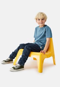 Sininen puuvillainen t-paita, siniset farkut ja vihreät kangastennarit, joita käyttää lapsi istumassa kirkkaan keltaisella muovituolilla.