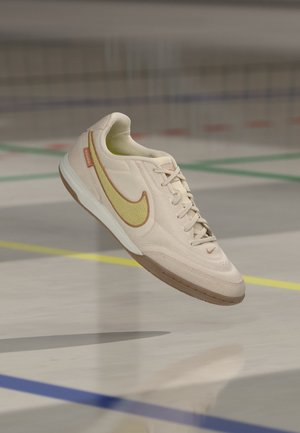 Μπεζ και κίτρινο αθλητικό παπούτσι Nike με κορδόνια που αιωρείται πάνω από ένα κλειστό αθλητικό γήπεδο με χρωματιστές διαχωριστικές γραμμές.