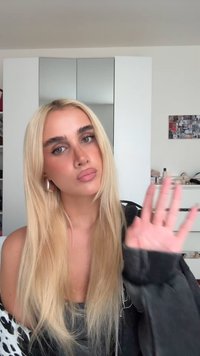 Blond włos opadający w subtelnych falach, ubrany w czarną bluzkę i ciemną kurtkę. Srebrne kolczyki w kształcie kół. Naturalny, neutralny makijaż. Tło to uporządkowany pokój.