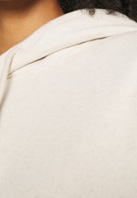 Sweat à capuche de couleur crème avec un tissu doux et texturé, doté d'un cordon de serrage au niveau du cou et d'une finition unie. Gros plan montrant les détails de la couture.