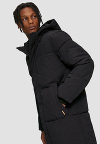 Schwarze Pufferjacke mit hoher Kapuze, gestepptem Design, Seitentaschen und einem orangefarbenen Akzent an der unteren Tasche. Strukturierter Stoff, mittellange Passform.