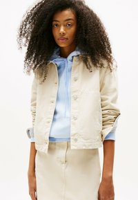 Chaqueta de algodón beige con botones a presión, llevada sobre una sudadera con capucha de color azul claro y una falda a juego, con mangas texturizadas y puños acanalados.