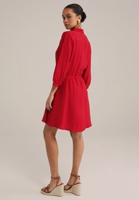 Robe rouge texturée avec un col, des manches bouffantes, une taille élastique et une jupe évasée. Associée à des sandales compensées beiges.