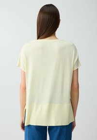 Femme aux cheveux bruns et lisses portant un haut ample jaune pâle à manches courtes et un jean bleu, de dos devant un fond clair uni.