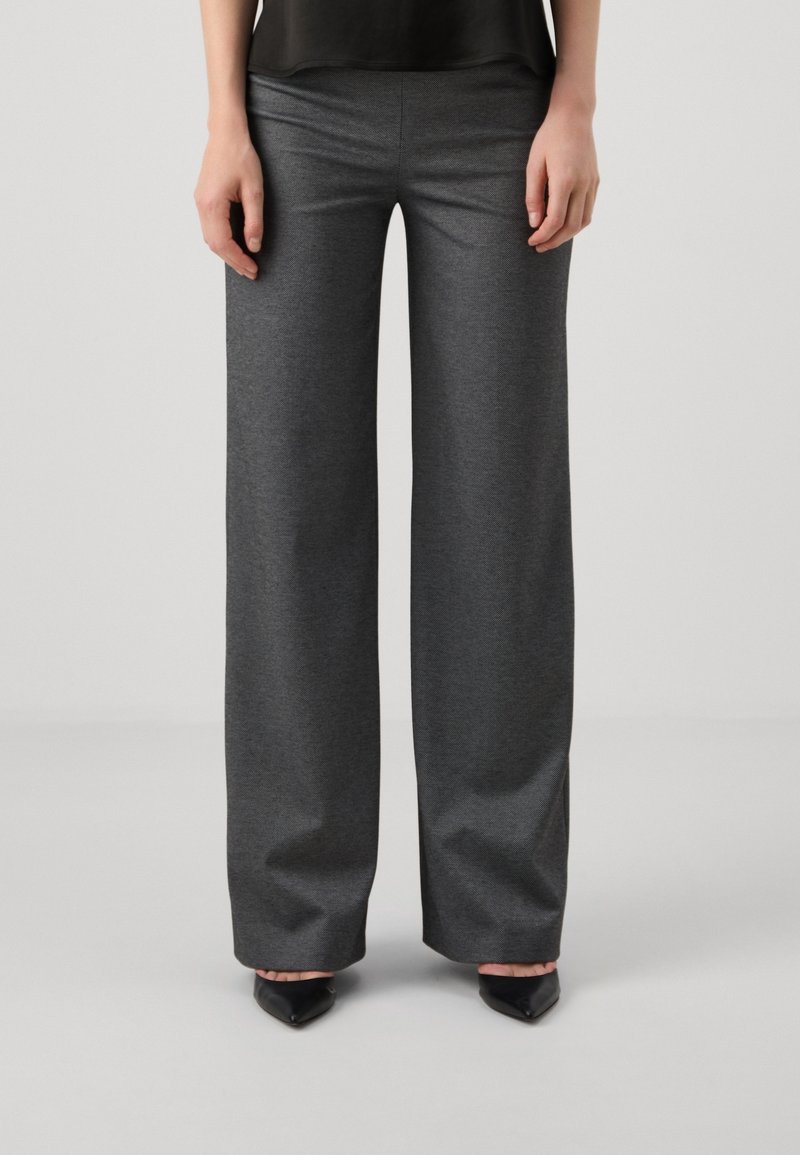 Femme portant un pantalon large gris foncé et des chaussures noires à bout pointu, debout devant un fond clair uni.