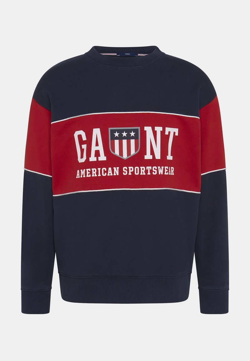 Sweatshirt bleu marine et rouge avec texte blanc "GAUNT" et "AMERICAN SPORTSWEAR" ainsi qu'un emblème en forme de bouclier avec des étoiles et des rayures sur la poitrine.