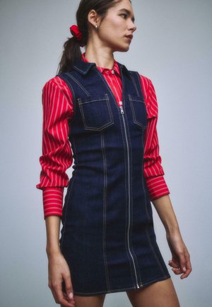 Frau trägt ein rotes, gestreiftes Hemd unter einem ärmellosen, dunkelblauen Jeanskleid mit Fronttaschen und Reißverschluss, Haare mit einem roten Haargummi gebunden.