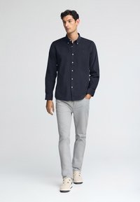 Camicia blu navy con colletto, abbinata a jeans grigi chiari e sneakers color crema. La camicia presenta un logo discreto sul petto.