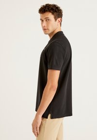 Černá polo košile s krátkými rukávy, límečkem a knoflíkovými akcenty. Tkání vypadá hladce, má klasický střih a boční rozparky pro komfort.