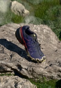 Sapatilha de trail running em azul escuro com acentos laranja, parte superior sintética texturizada, fecho de cadarços, sola cushioned preta e padrão de rasto agressivo.