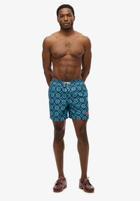Zwemshorts in het blauw met een geometrisch patroon. Elastische tailleband met trekkoord. Model draagt bruine veterschoenen.