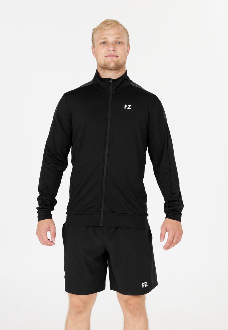 En blond man som bär en svart zip-up-jacka och svarta shorts med "FZ"-logotyper, stående med avslappnade armar mot en plain vit bakgrund.