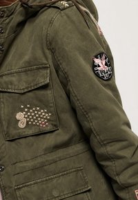 Olijfgroene jas met capuchon, voorzien van twee voorzakken, geborduurde accenten en een patch op de mouw. Gestructureerde stof met een ontspannen pasvorm.