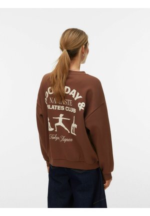 Donna con coda di cavallo che indossa una felpa marrone con pose bianche di yoga e pilates e testo che recita "Good Day & Namaste Pilates Club Tokyo, Japan."
