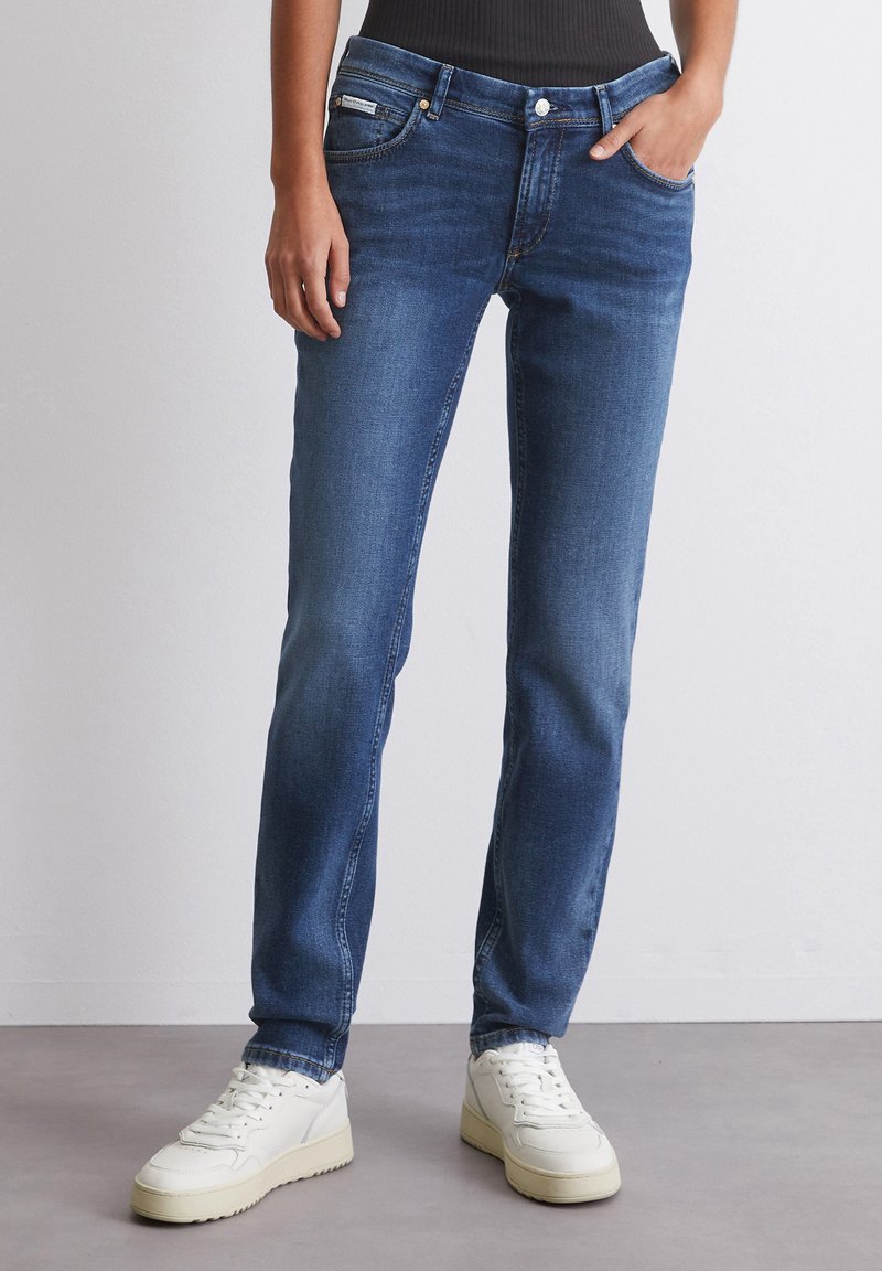 Tynne mørkeblå denimjeans med fem lommer, sømdetaljer og sølvfarget hardware, kombinert med hvite sneakers.