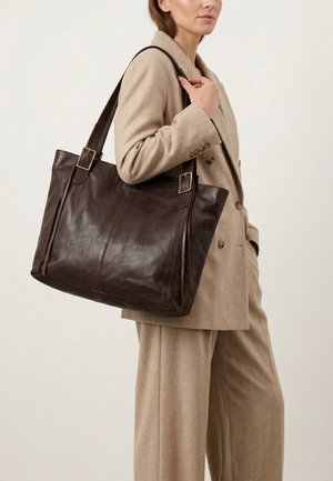 Vrouw in beige pak draagt een grote donkerbruine leren tas over de schouder tegen een effen achtergrond.