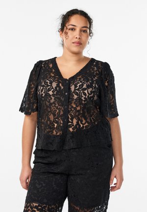 Kobieta o kręconych włosach nosi czarną koronkową bluzkę na guziki z kwiatowym wzorem oraz pasujące szorty na białym tle.