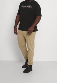 Černé tričko s krátkým rukávem s bílým logem a khaki kalhoty s vyhrnutými nohavicemi. Černé kotníkové boty s texturovanou podrážkou doplňují outfit.