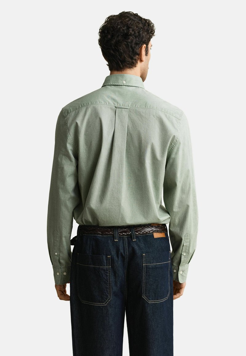 Hombre con camisa de botones verde claro y vaqueros azul oscuro, de pie con la espalda hacia adelante sobre un fondo liso.