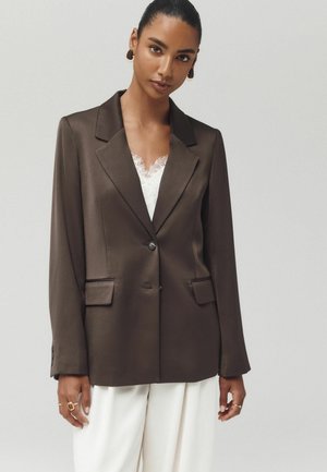 Kvinde iført en mørkebrun blazer over en hvid blondebluse og hvide bukser, stående mod en ensfarvet lys baggrund.