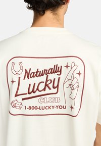 Cream cotton t-shirt featuring red graphic with "Naturally Lucky" text, a hand gesture, dice, and a horseshoe design. Short sleeves.