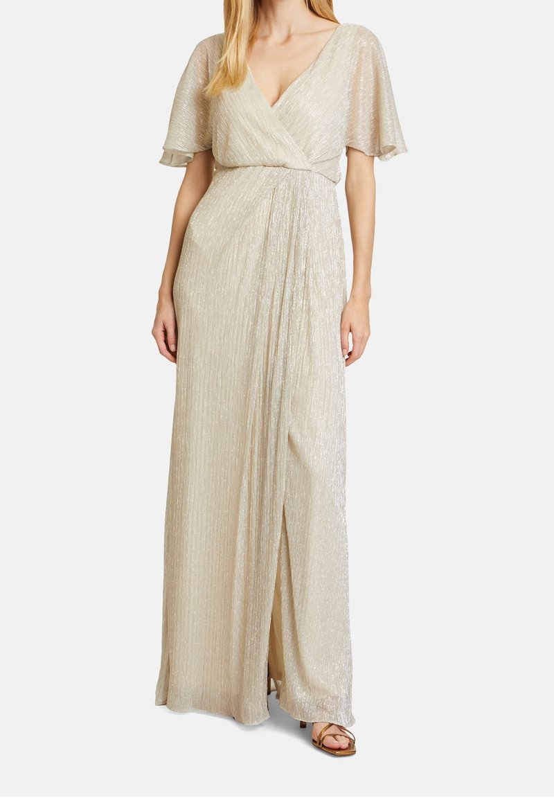 Femme portant une robe champagne longue au sol, scintillante, à manches papillon, décolleté en V et devant drapé, debout devant un fond uni.