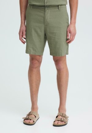 Mand iført olivengrønne knapshorts og beige sandaler med dobbelt rem, stående på en ensfarvet lys baggrund.