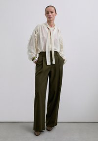 Mujer de pie con una blusa suelta color crema con volantes, pantalones anchos color oliva, zapatos marrones de punta y pulseras de plata en su muñeca.