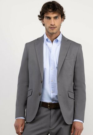 Hombre con chaqueta de traje gris, camisa azul clara y cinturón marrón, de pie frente a un fondo blanco liso.