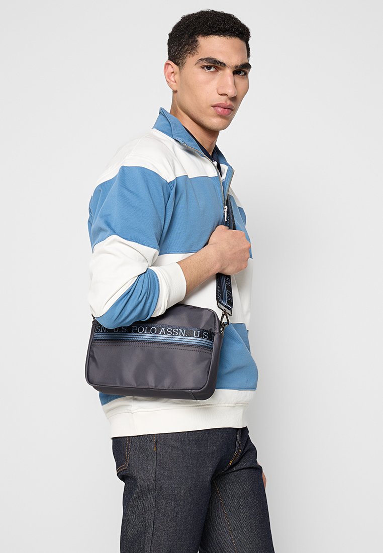 Ciemnoszara nylonowa torba na ramię z niebieskim paskiem z logo. Charakteryzuje się prostokątnym kształtem i gładką fakturą. Posiada uchwyt do noszenia.