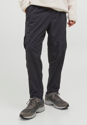 Personne portant un pantalon cargo noir et des baskets grises, debout contre un fond clair uni, corps inférieur visible.