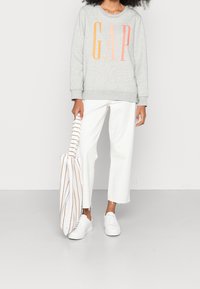 Felpa grigia con logo "GAP" in arancione e rosa, jeans bianchi a vita corta con orlo sfrangiato e sneakers bianche. Borsa a strisce in mano.