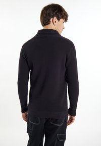 Maglione nero a maglia testurizzata con colletto alto, maniche lunghe e polsini a coste; indossato da un modello maschile di spalle rispetto alla macchina fotografica.