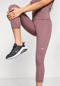 Melnās un baltās Nike sporta kedas ar acs augšdaļu; modele ģērbusies pieguļošās, līdz ceļiem garās, gaiši rozīgās legingās ar šuvju detaļām.