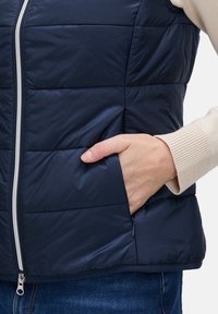 Marinefarbener gepolsterter Weste mit Reißverschluss, versehen mit einer Seitentasche und einem gerippten beigen Bündchen. Glatte Textur mit gesteppten Abschnitten.