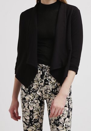 Kvinde iført en sort cardigan med åben front over en sort top, kombineret med højtaljede bukser med blomsterprint i beige, sort og hvide nuancer.