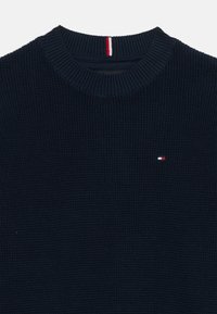 Pull en laine navy avec une finition texturée, présentant un col rond côtelé et un petit logo en rouge, blanc et bleu sur la poitrine.