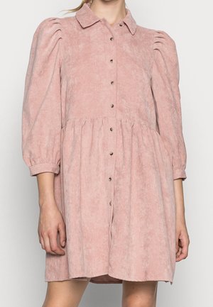 Różowa sukienka sztruksowa z bufkami na długich rękawach, zapinana na guziki, z kołnierzykiem i marszczonym pasem, noszona przez stojącą osobę.
