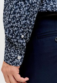 Mehe käsi tumesiniste pükste tasku kõrval, kannab pika varrukaga särki, mille mansetid on tumesinise ja valge lillemustriga ning valgete nuppudega.