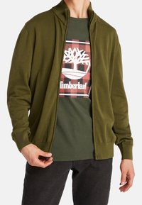 Cardigan olive verde con cierre y puños acanalados, combinado con una camiseta verde oscura que presenta un logotipo blanco de Timberland sobre un fondo de cuadros.
