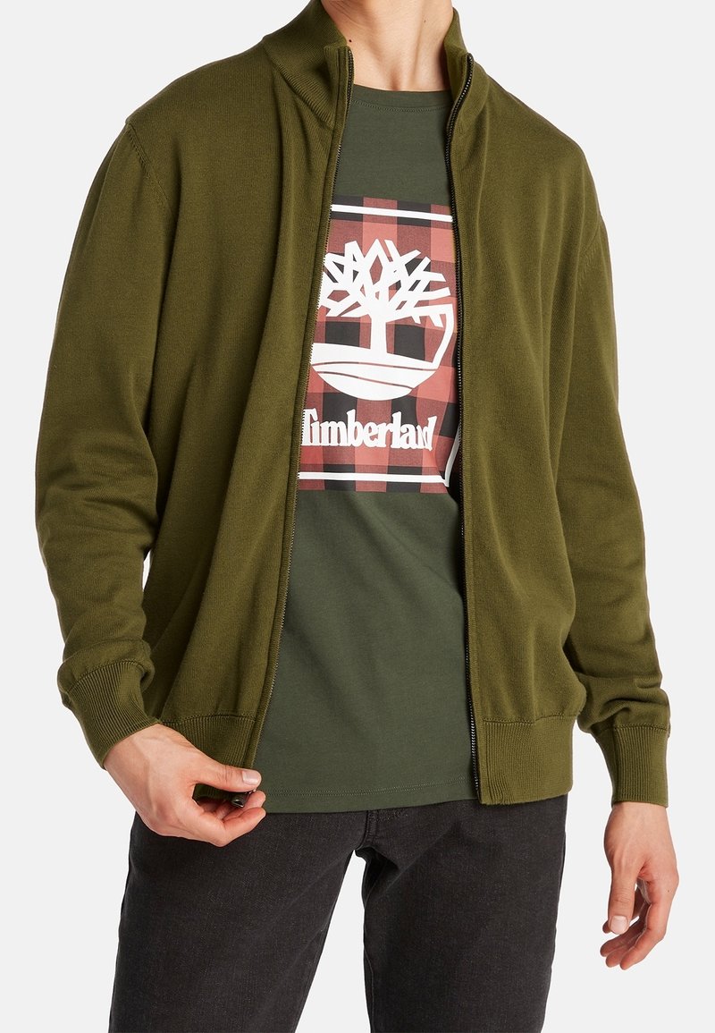 Olivgrüner Reißverschluss-Pullover mit Rippbündchen, kombiniert mit einem dunkelgrünen T-Shirt, das ein weißes Timberland-Logo auf einem karierten Hintergrund zeigt.